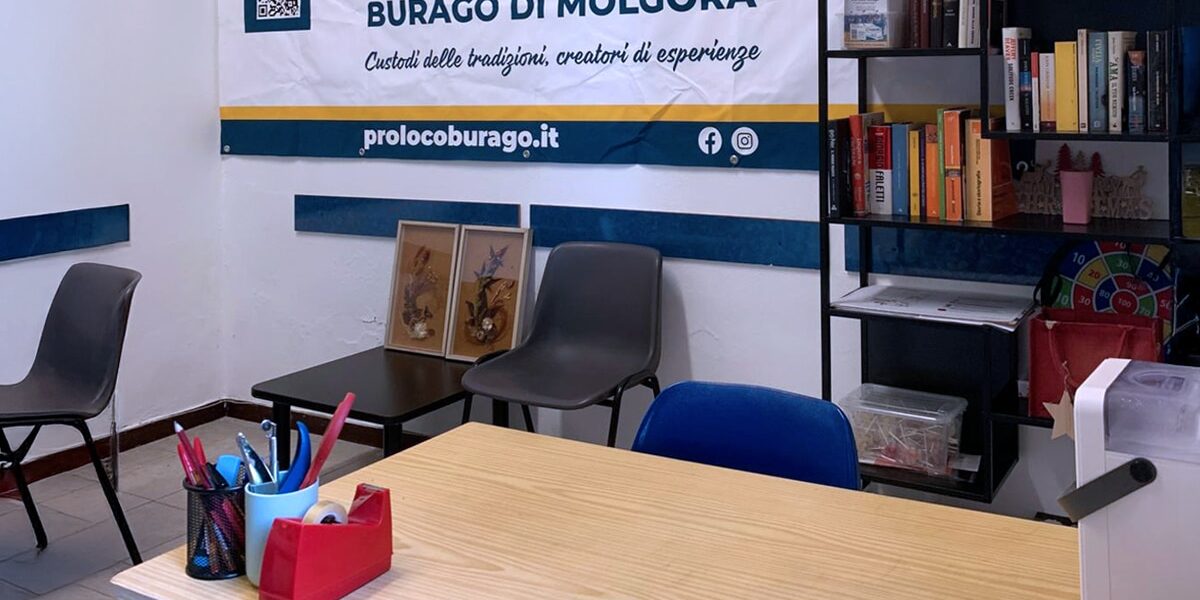 Pro Loco Presente – apertura sede il lunedì sera Interno della sede Pro Loco Burago con tavolo, sedie, libreria e banner dell’associazione appeso alla parete.